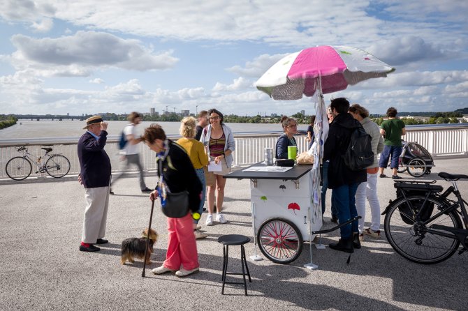 Plusieurs personnes autour d'un petit comptoire à roues surmonté d'un parasol blanc et rose sur le pont Simone-Veil